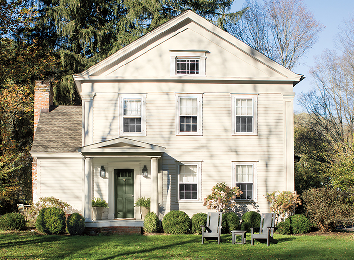 A white-painted home exterior with a dark green front door, a covered entryway with pillars, and a lush green lawn with two green chairs.