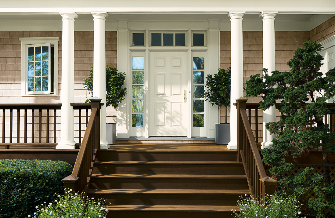 A home exterior front porch with a white-painted front door and columns, taupe stained shingles, and brown stairs and railings.
